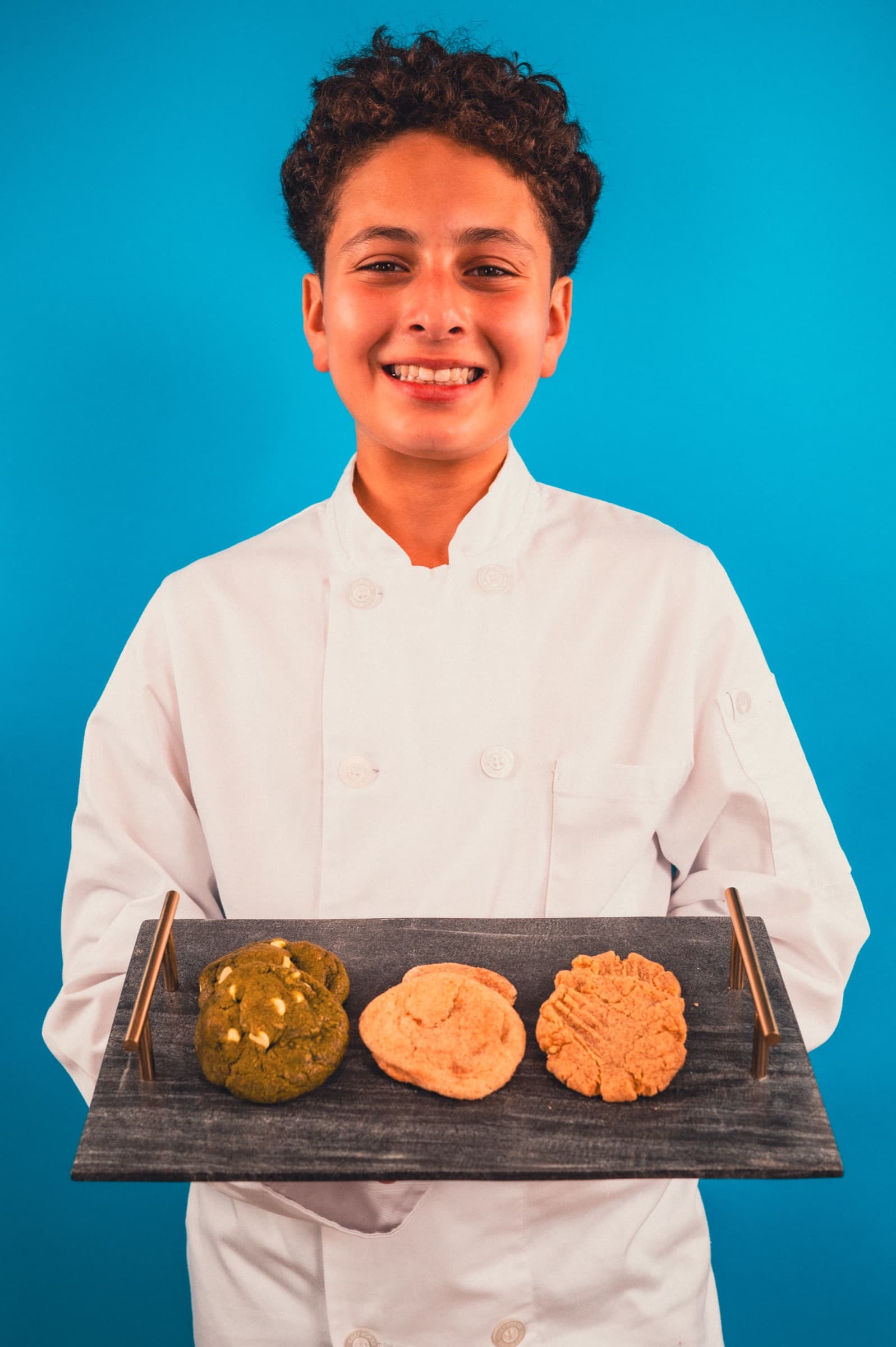 Nour in a chef coat holding a tray of matcha, sugar, and peanut butter cookies